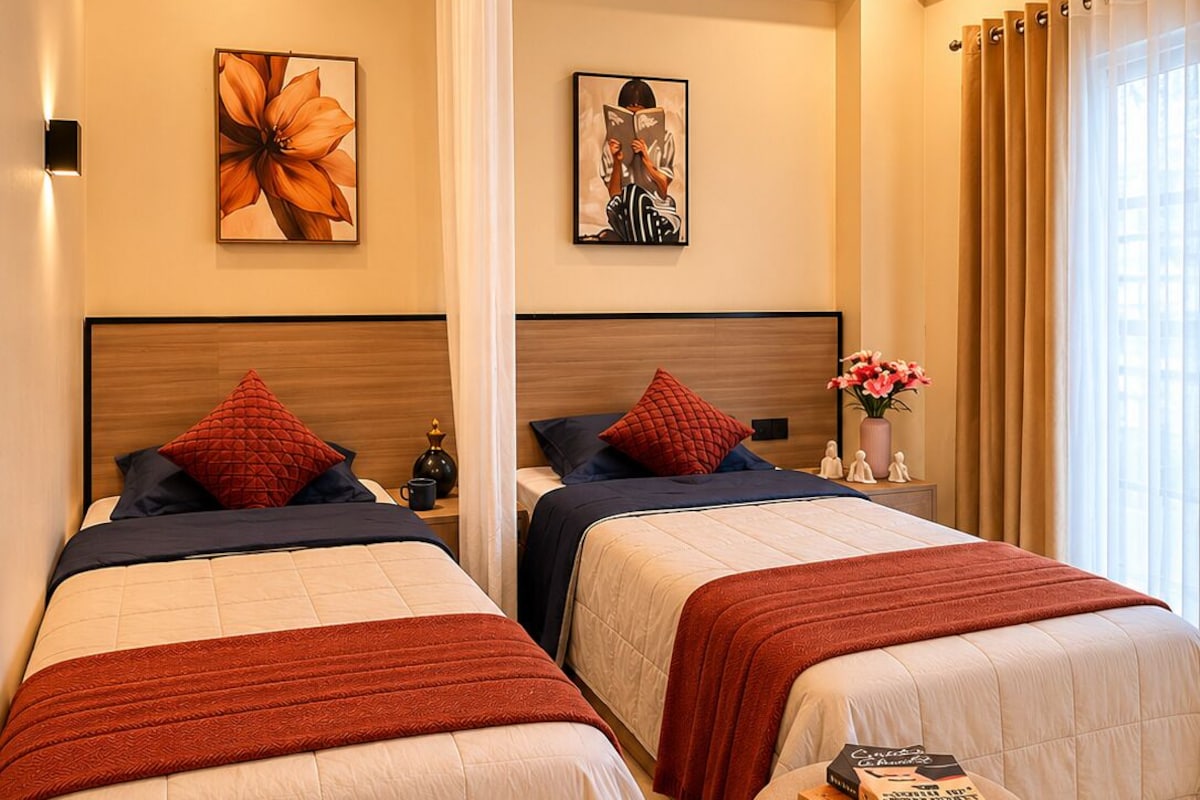 Une chambre d’hôtel avec deux lits, une table de chevet ornée d’un vase de fleurs et une grande fenêtre avec des rideaux.