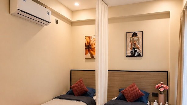 Una habitación de hotel con dos camas, un otoman redondo, una mesita de noche con un florero y obras de arte en las paredes.