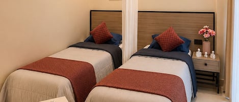 A hotel room with two beds, a round ottoman, a nightstand with a vase, and artwork on the walls.