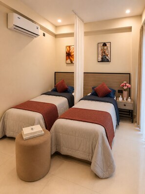 A hotel room with two beds, a round ottoman, a nightstand with a vase, and artwork on the walls. - C Suites Maclean (Bengaluru)