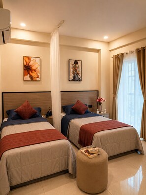 Habitación de hotel con dos camas, una mesita de noche, un otoman redondo y obras de arte en las paredes.