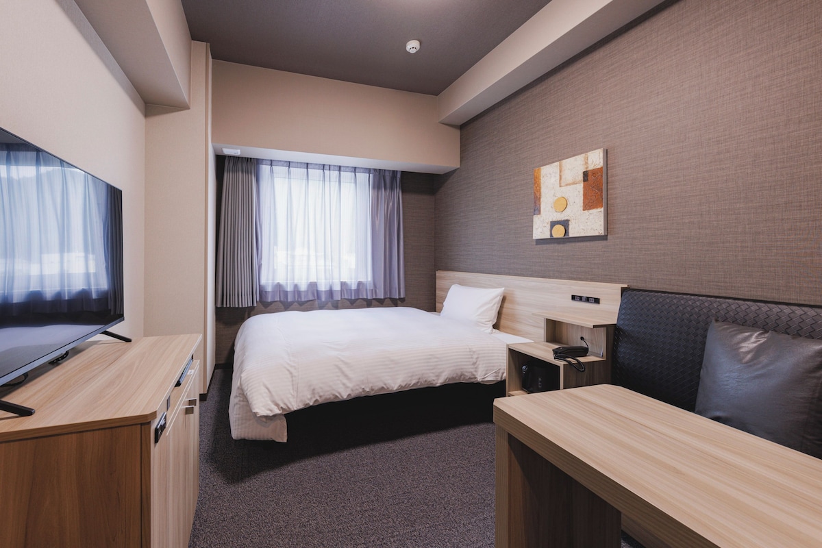 A hotel room with a bed, a desk, a television, and a painting on the wall.
