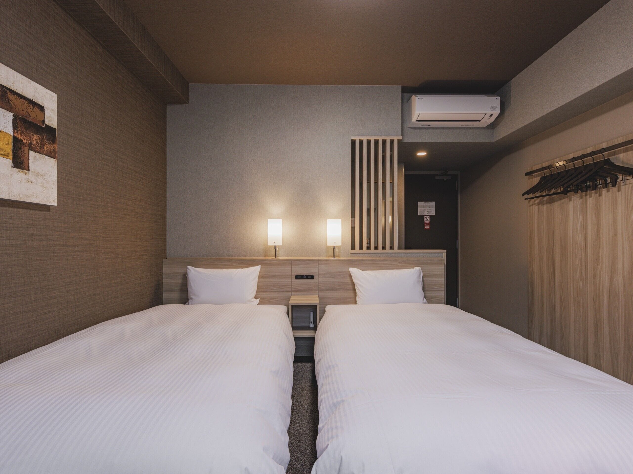 Una habitación de hotel con dos camas individuales, aire acondicionado de pared y una mesita de noche pequeña con lámpara.