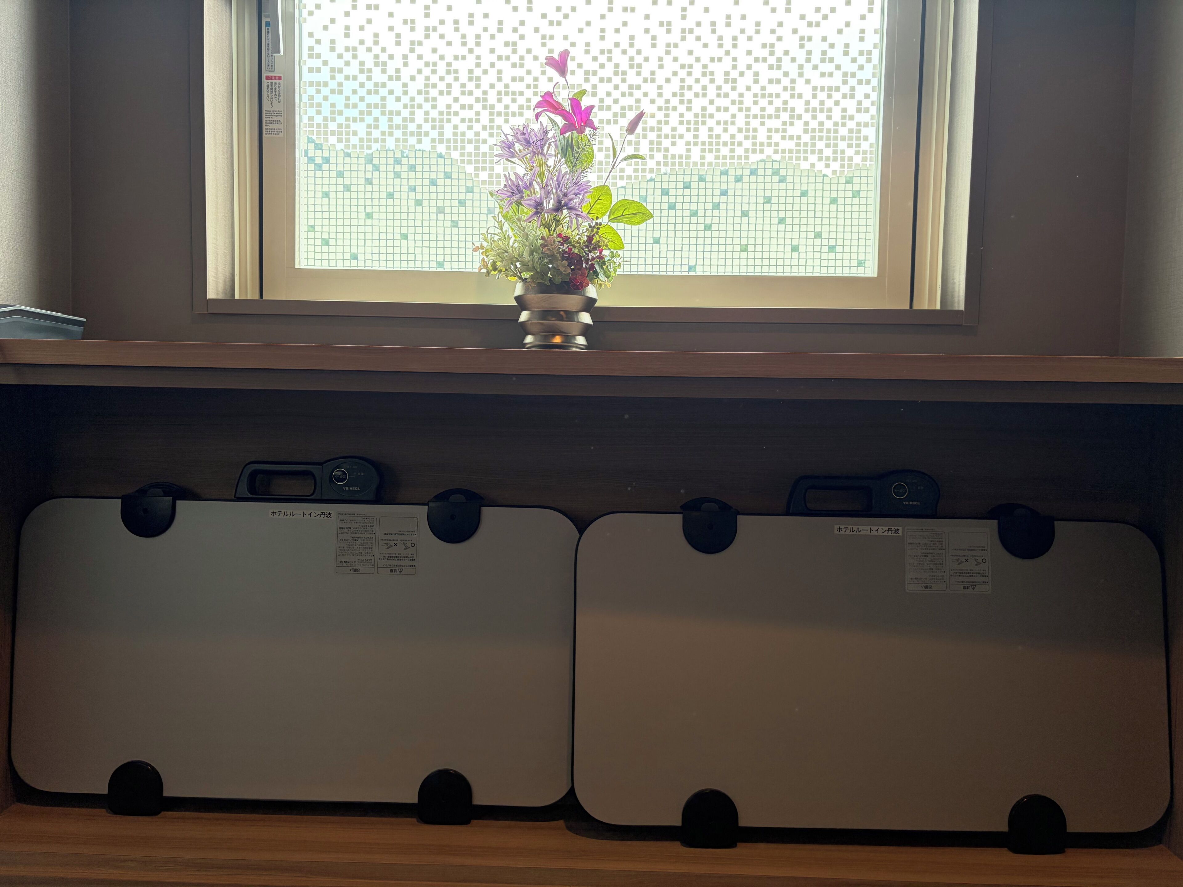 A window with a view of a fence, a potted plant with flowers, and two suitcases on a shelf below.