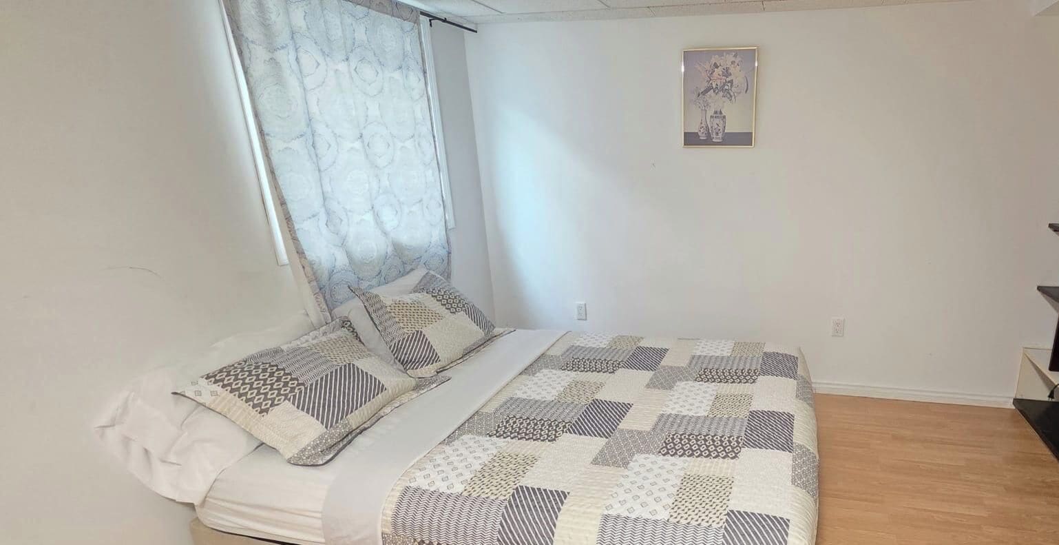 Quarto com cama, janela com cortina estampada, quadro na parede e piso de madeira.