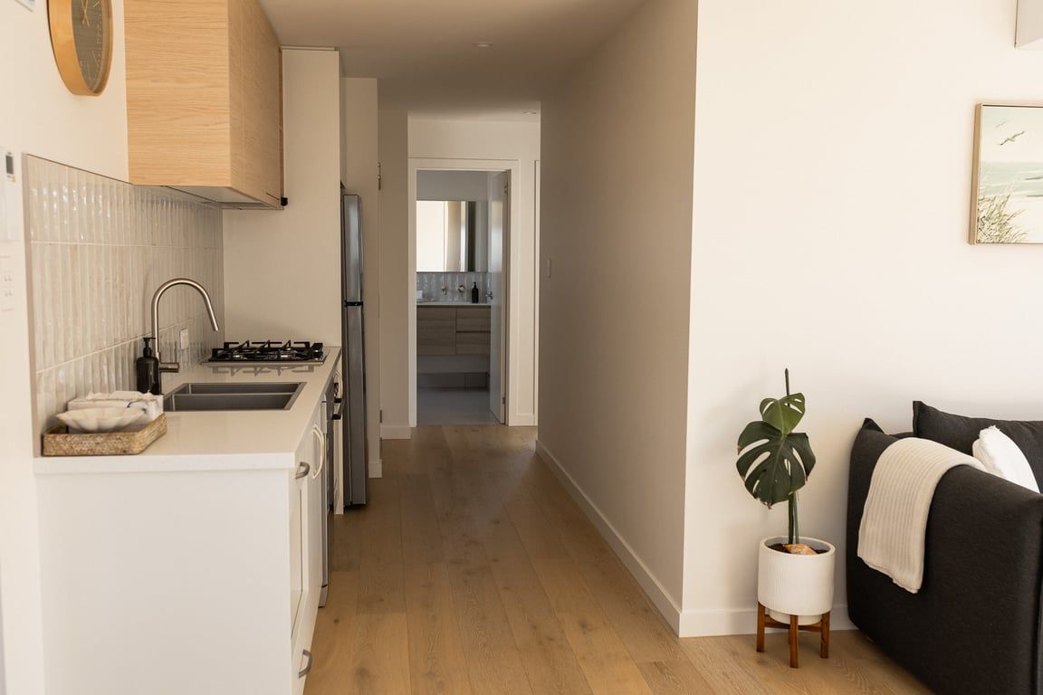Ett modernt kök med en diskho i rostfritt stål, en spis och ett kylskåp. En hall leder vidare till en matplats och ett vardagsrum med en svart soffa och en krukväxt.