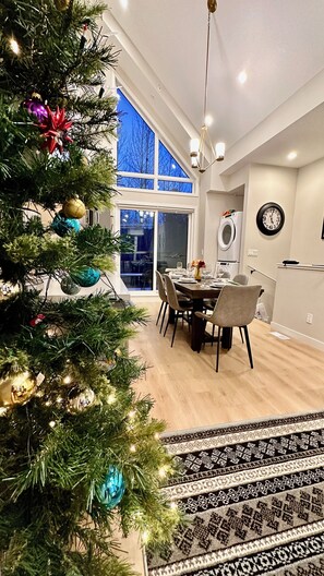 A modern dining area with a Christmas tree, a dining table set for four, and a large window with a view. - The Canmore Mountain Twin Retreat with 4BR • 2 Homes • Heated Pool & Hot Tub (Canmore)