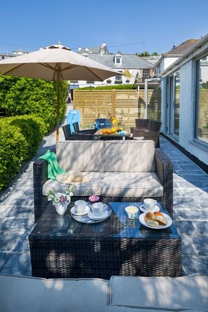 Uma área externa com assentos de vime, uma mesa com xícaras e doces, e um grande guarda-sol para dar sombra.