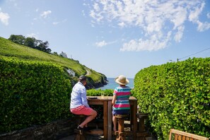 Zwei Personen sitzen an einem Holztisch mit Blick auf das Meer und einen grünen Hügel.