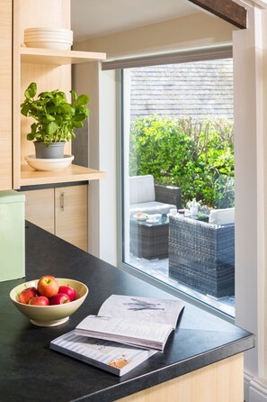 Una cocina con vista por la ventana a muebles de exterior, un bol de manzanas y una revista.