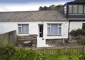 A white cottage with a stone foundation, a wooden door, and a small patio with a table and chairs. - Rose Cottage - sleeps 4 guests  in 2 bedrooms (Port Gaverne)