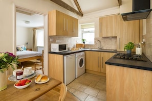 A compact kitchen with wooden cabinets, a microwave, a washing machine, and a dining area with a table set for breakfast. - Rose Cottage - sleeps 4 guests  in 2 bedrooms (Port Gaverne)