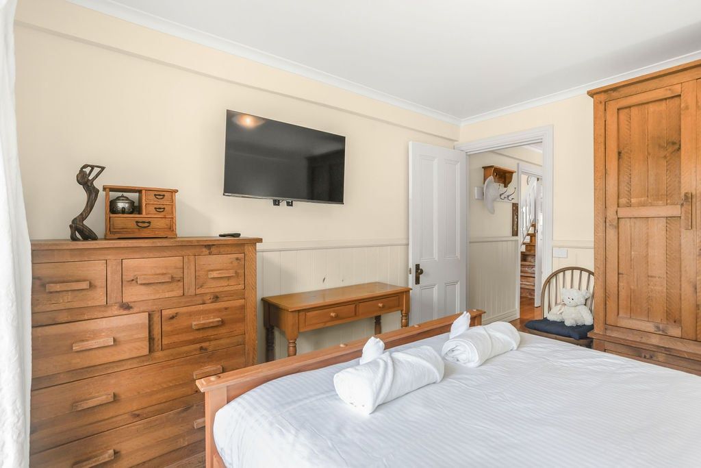 Ein Schlafzimmer mit einem Holzkoffer, einem Nachttisch, einem Bett mit weißer Bettwäsche, einem an der Wand befestigten Fernseher und einem Holzschrank.
