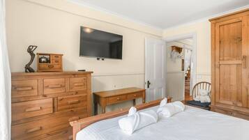 Ein Schlafzimmer mit einem Holzkoffer, einem Nachttisch, einem Bett mit weißer Bettwäsche, einem an der Wand befestigten Fernseher und einem Holzschrank.