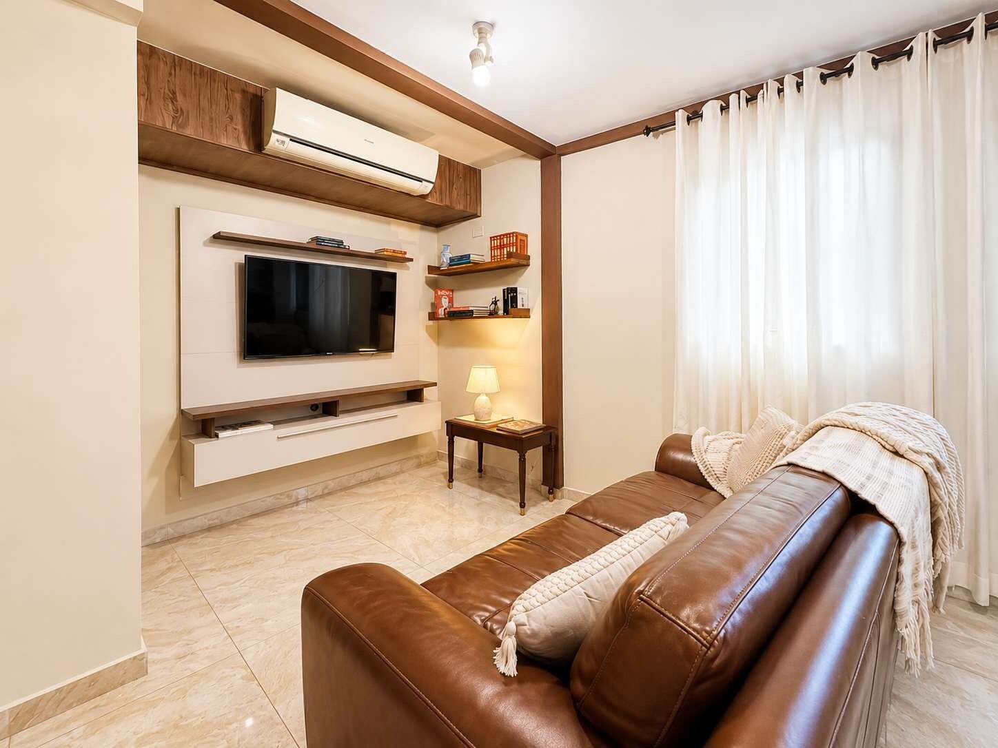 A living room with a brown leather sofa, a flat-screen TV, a small table with a lamp, and a bookshelf.