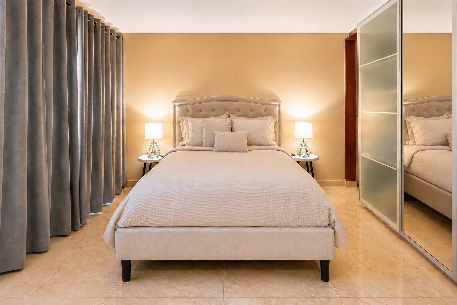 Habitación de hotel con una cama grande, dos mesitas de noche con lámparas, un armario con espejo y un ventanal cubierto con cortinas.