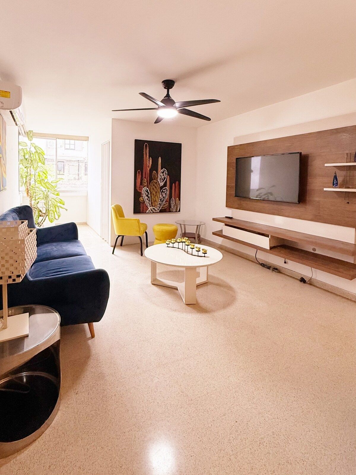A modern living room with a blue sofa, yellow chairs, a coffee table, a ceiling fan, and a flat-screen TV mounted on a wooden wall.