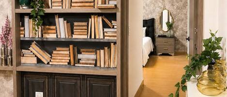 A room with a large bookshelf filled with books, a bedside table with a book, a mirror, and a potted plant.