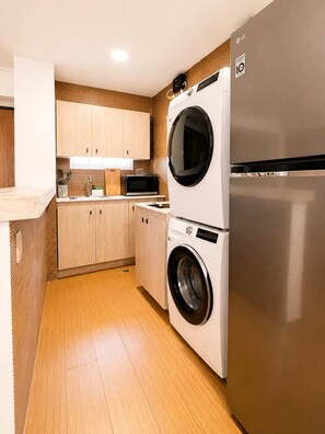 A compact kitchen with wooden cabinets, a washing machine, a microwave, and a stainless steel refrigerator. - Skyline Views Casco Viejo Apt | Historic (Panamá)