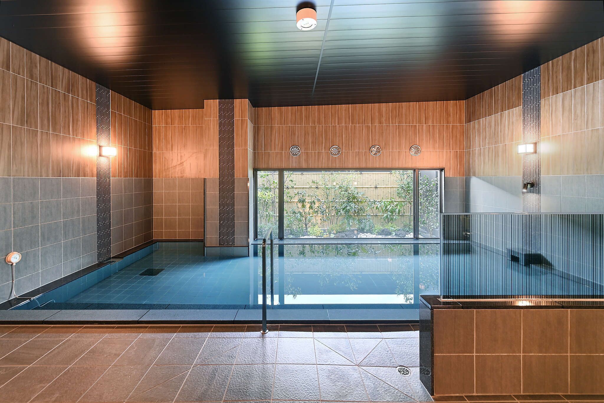 A modern indoor swimming pool with a glass barrier, wooden panel walls, and a view of greenery outside.