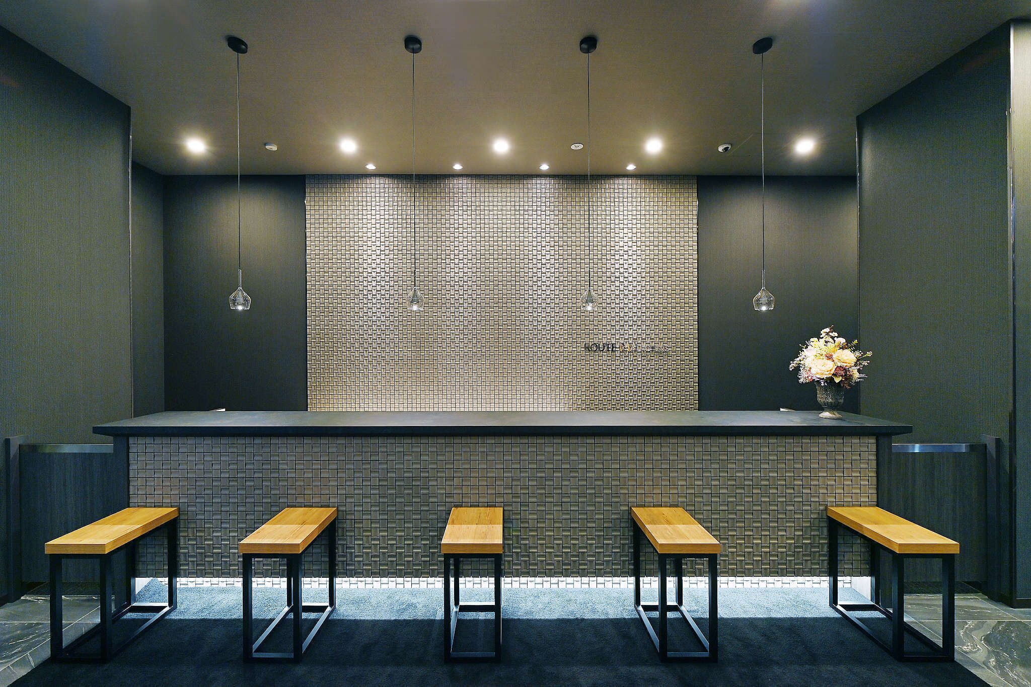 A bar area with a tiled backsplash, five wooden stools, hanging pendant lights, and a vase with flowers on the counter.