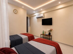A hotel room with two beds, a TV, an air conditioning unit, and a small table with a vase and books. - C Suites Shelton (Bengaluru)