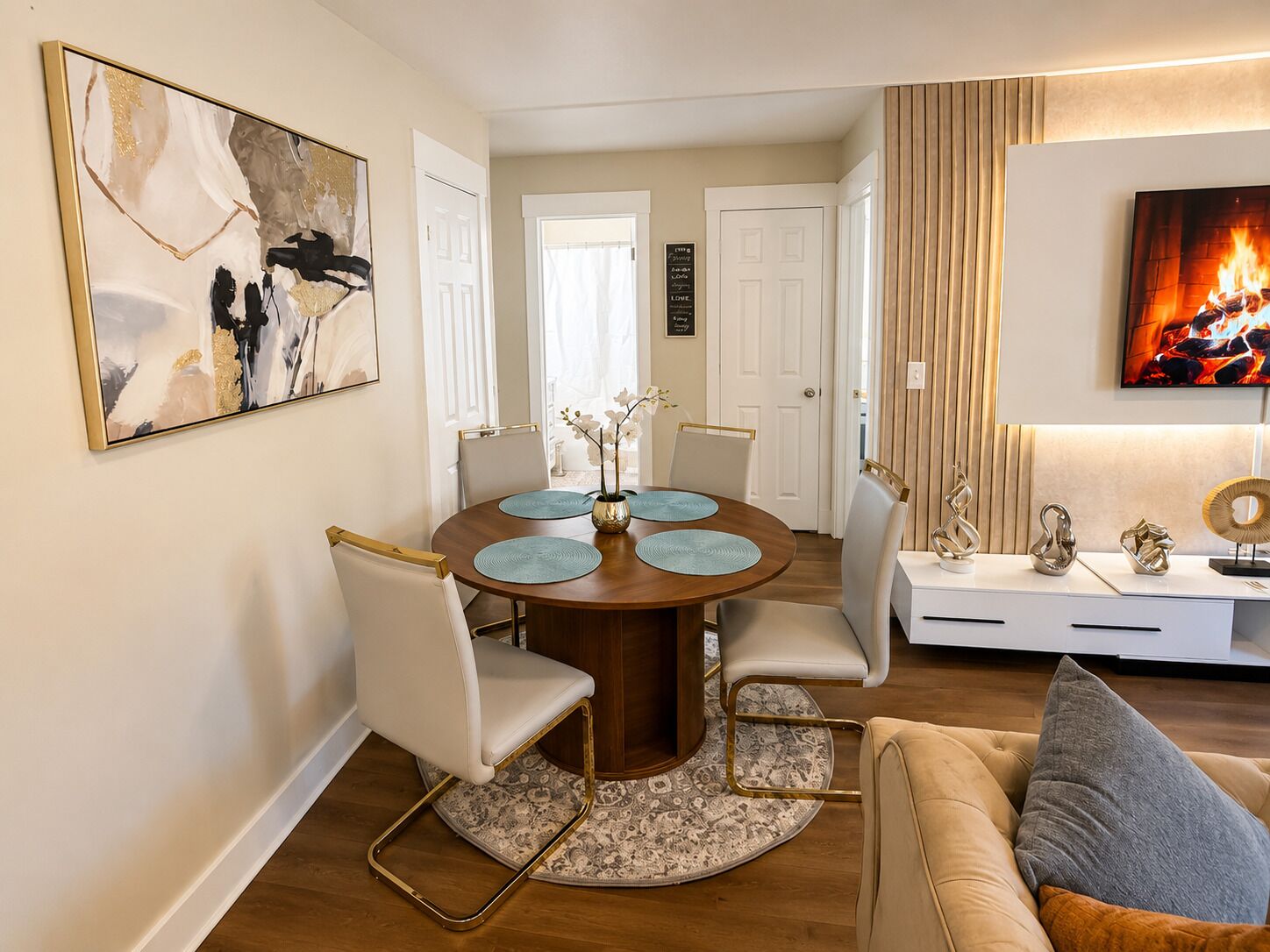 Un coin repas avec une table ronde, des chaises et un tapis décoratif. Un téléviseur est fixé au mur et un canapé se trouve au fond de la pièce.
