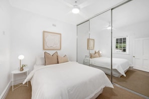 Ein Schlafzimmer mit einem großen Spiegel, einem Bett mit weißer Bettwäsche, einem kleinen Nachttisch mit Lampe und einem Fenster mit Blick auf Grünflächen.