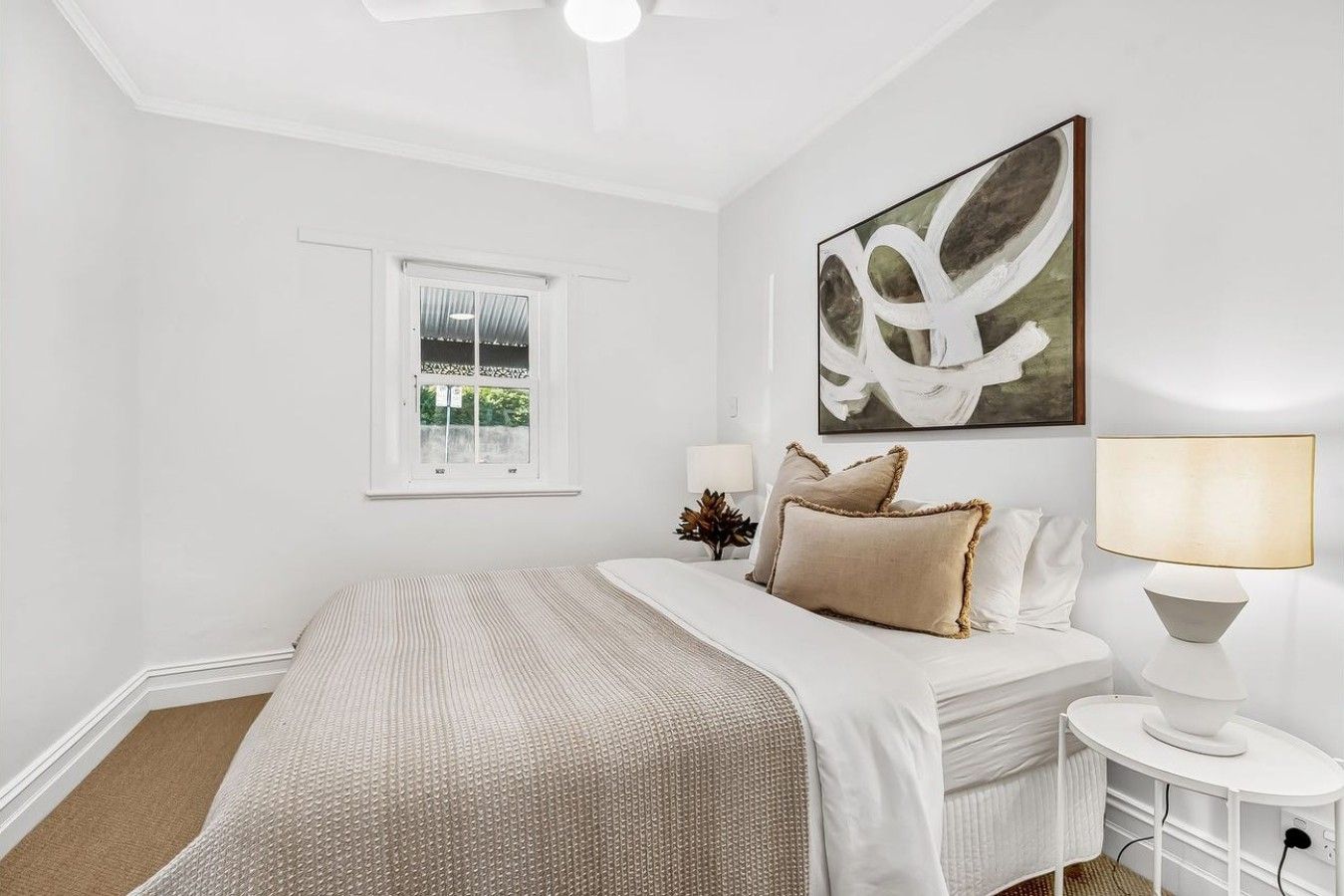 A bedroom with a bed, a window, a wall-mounted artwork, and a bedside table with a lamp.