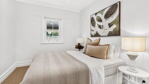 A bedroom with a bed, a window, a wall-mounted artwork, and a bedside table with a lamp. - Renovated character cottage, walk to Melbourne St and the city (North Adelaide)