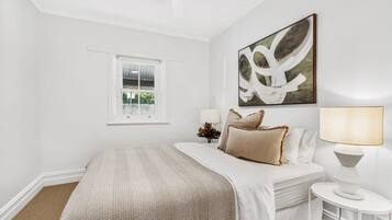 A bedroom with a bed, a window, a wall-mounted artwork, and a bedside table with a lamp.