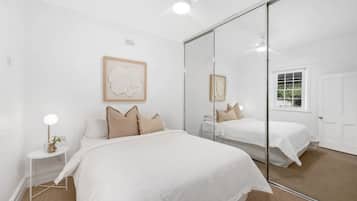 Une chambre avec un grand miroir, un lit recouvert de draps blancs, une petite table de chevet avec une lampe et une fenêtre donnant sur un paysage verdoyant.