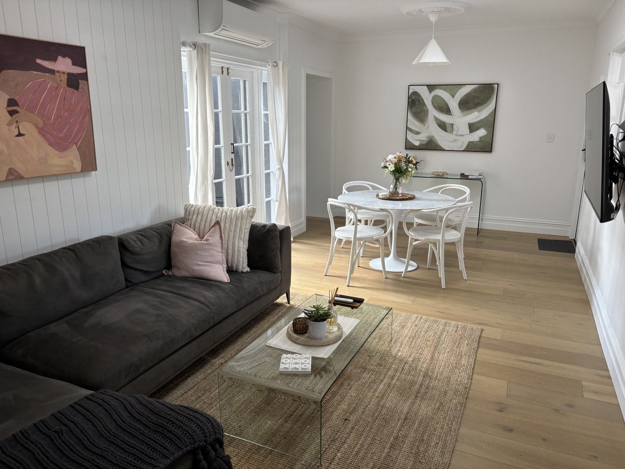Un salon moderne avec un canapé gris foncé, une table basse agrémentée d’objets décoratifs, un coin repas avec des chaises blanches et un grand tableau abstrait accroché au mur.
