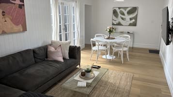 Un salon moderne avec un canapé gris foncé, une table basse agrémentée d’objets décoratifs, un coin repas avec des chaises blanches et un grand tableau abstrait accroché au mur.
