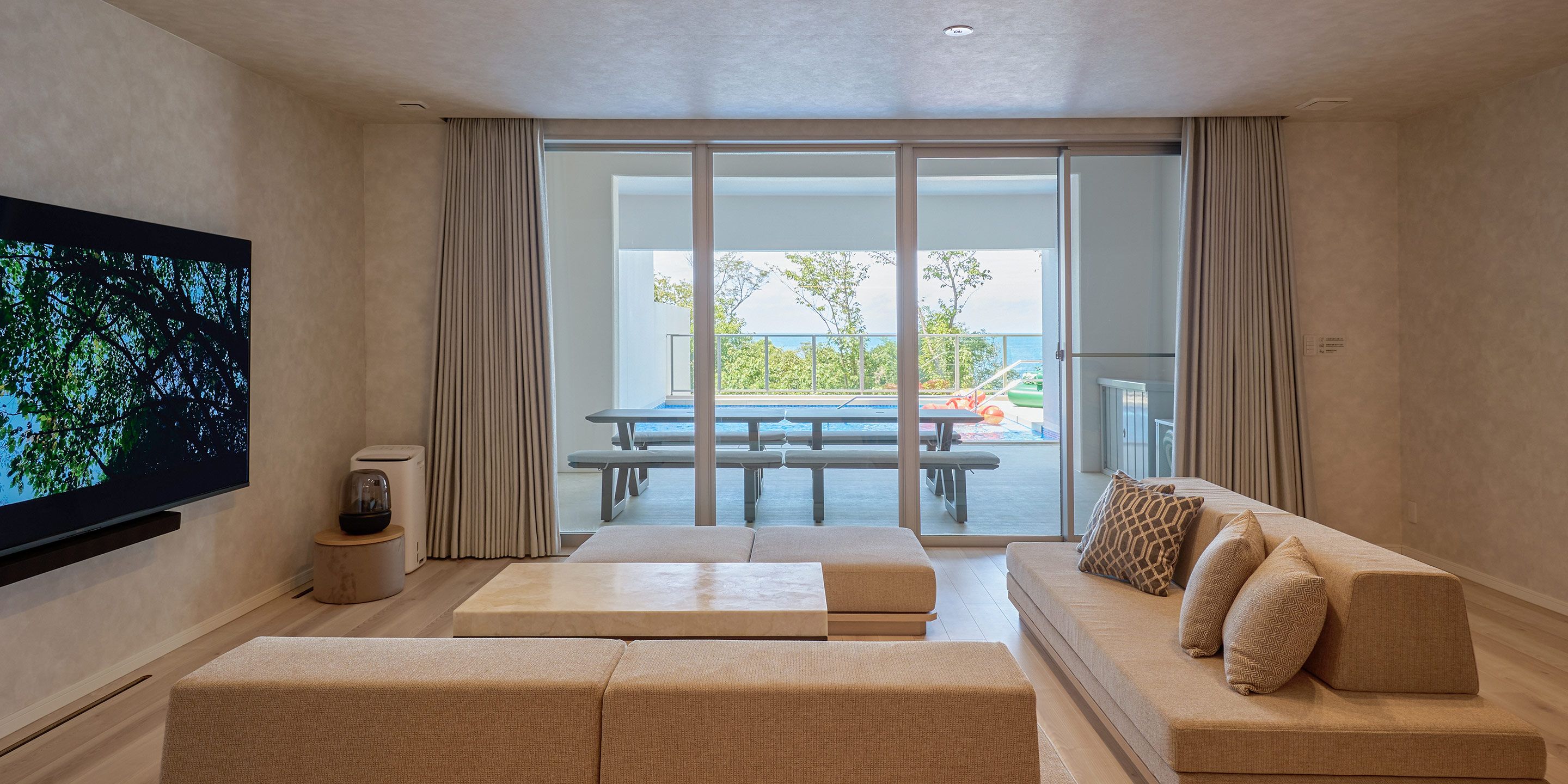 A modern living room with a flat-screen TV, a sofa, a coffee table, and a view of a pool and balcony.