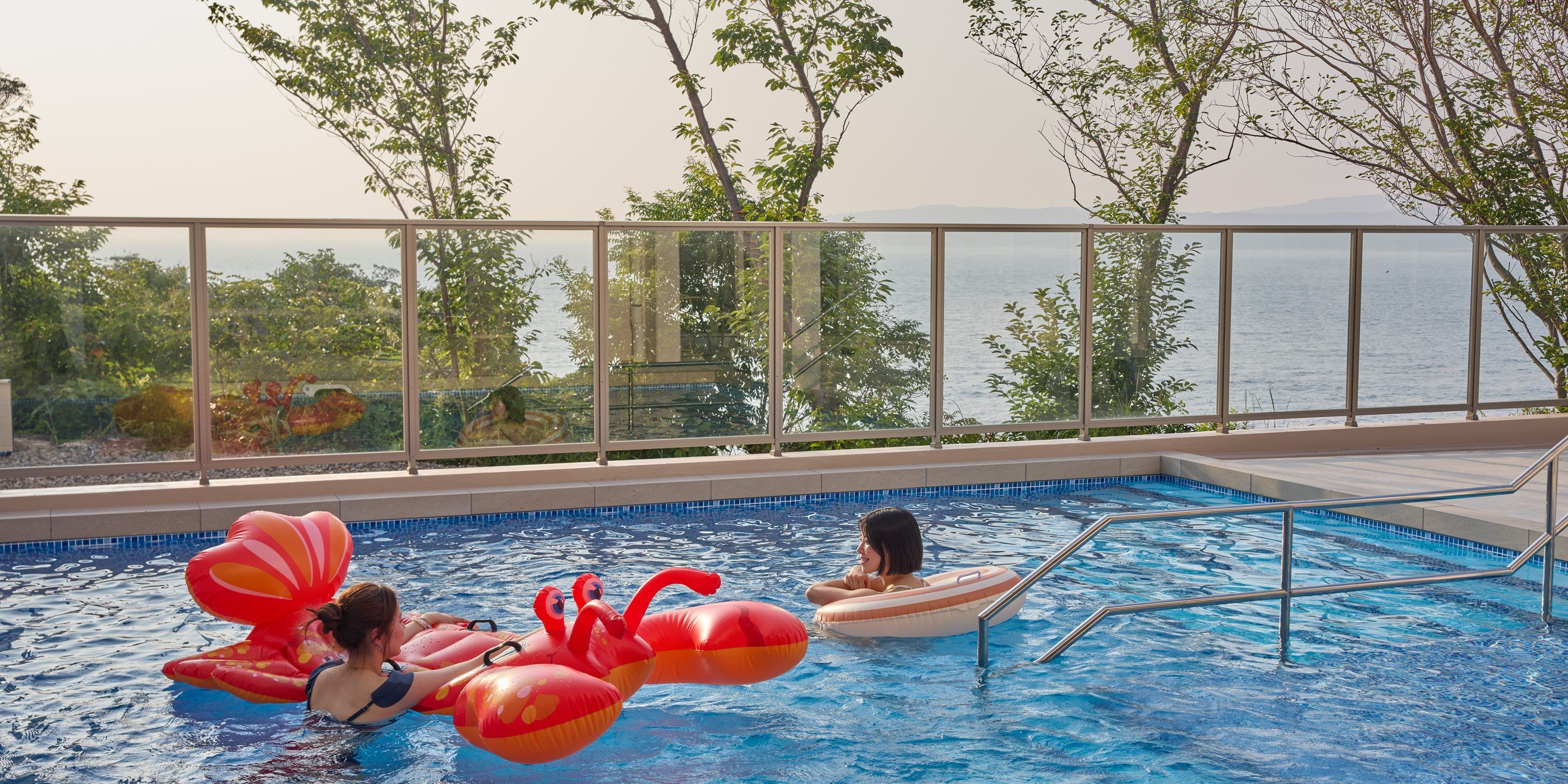 Piscina com vista para o mar, cercada por guarda-vidros de vidro e árvores.