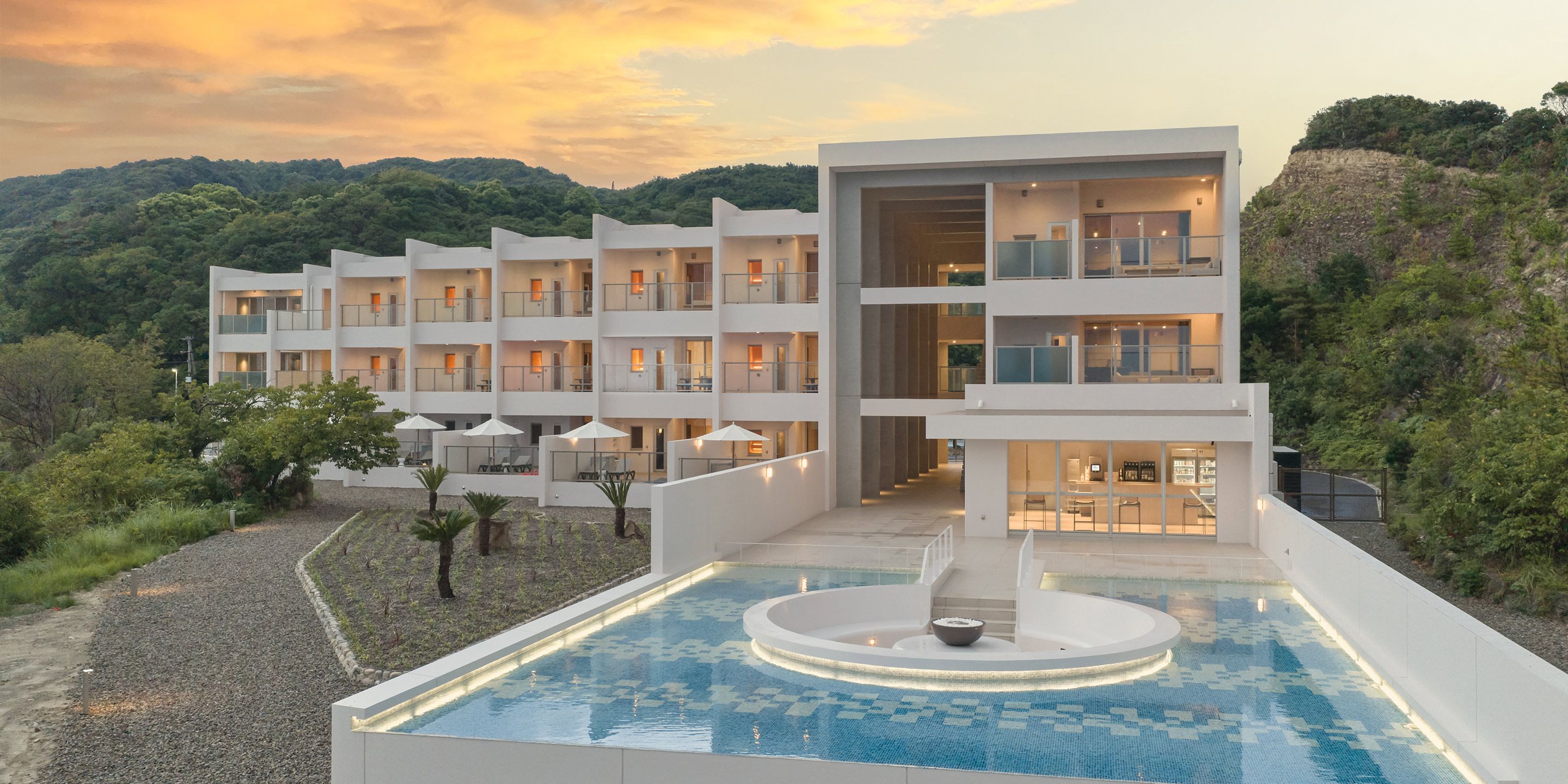 A modern hotel with multiple balconies, a swimming pool, and a view of the surrounding hills.