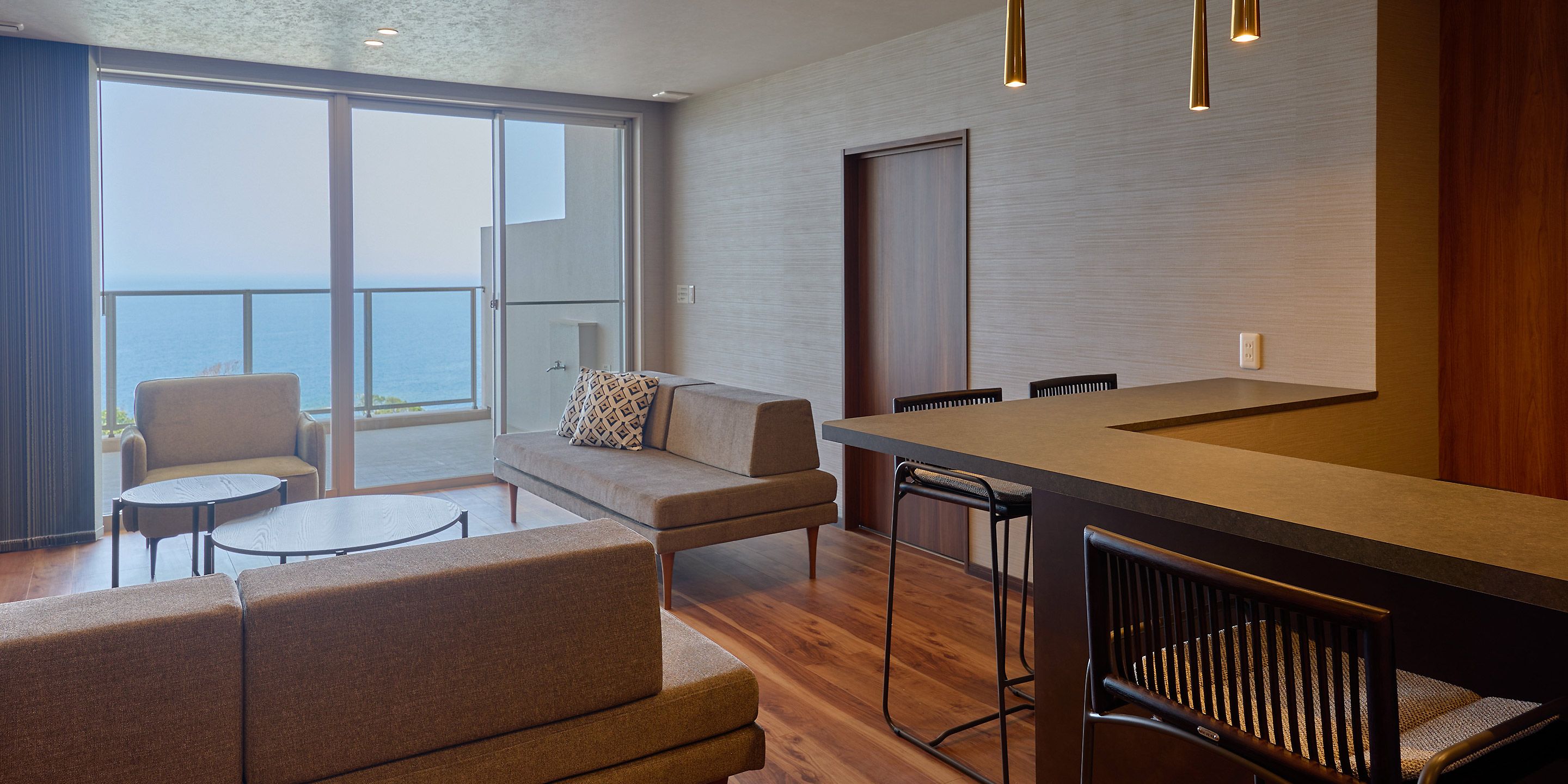 A modern hotel room with a balcony, a sofa, a dining table, and bar stools.