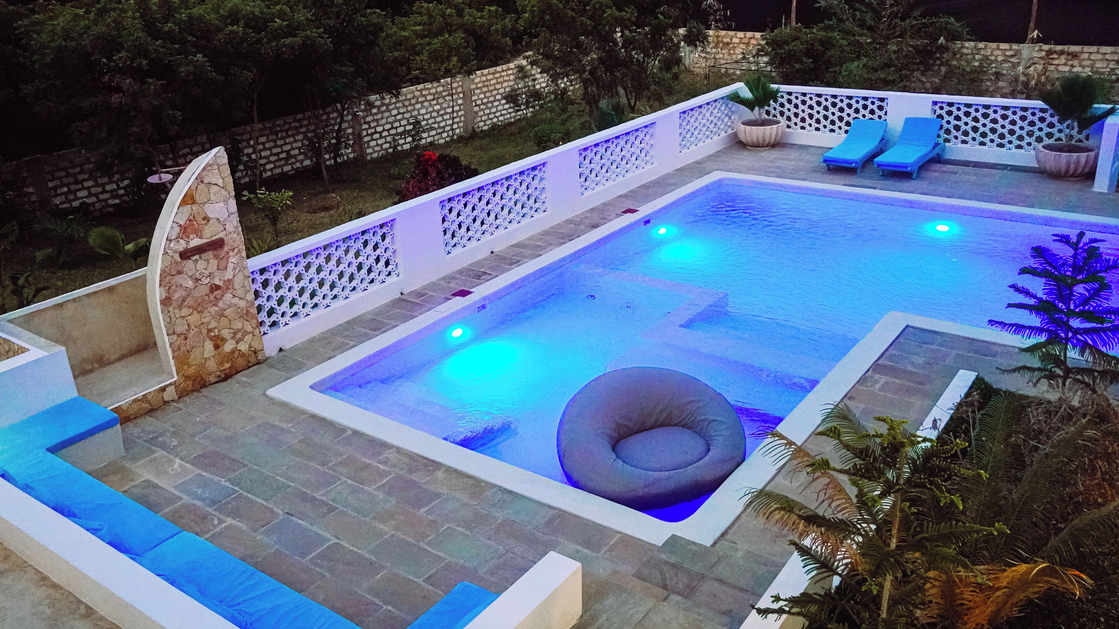 A rectangular swimming pool with a white border, a lounge area with blue cushions, and a stone wall with a decorative pattern.