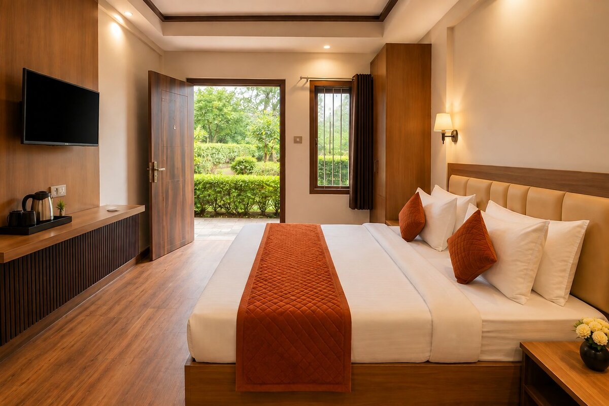 A hotel room with a bed, a TV, a bedside table, a window with a view of greenery, and a door leading to an outdoor area.