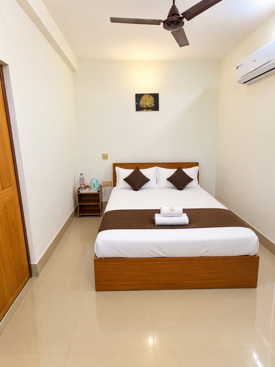 A hotel room with a bed, a ceiling fan, an air conditioning unit, a nightstand, and a wall-mounted artwork.