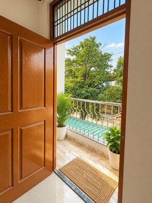 Una puerta de madera que da a un balcón con barandilla metálica, plantas en macetas y vistas despejadas a árboles y edificios.
