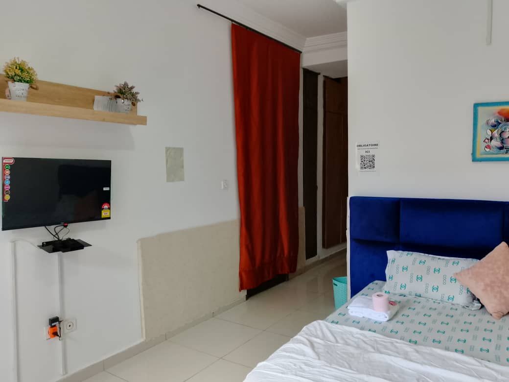 A hotel room with a bed, a television, a shelf with small plants, a red curtain, and a sign on the wall.
