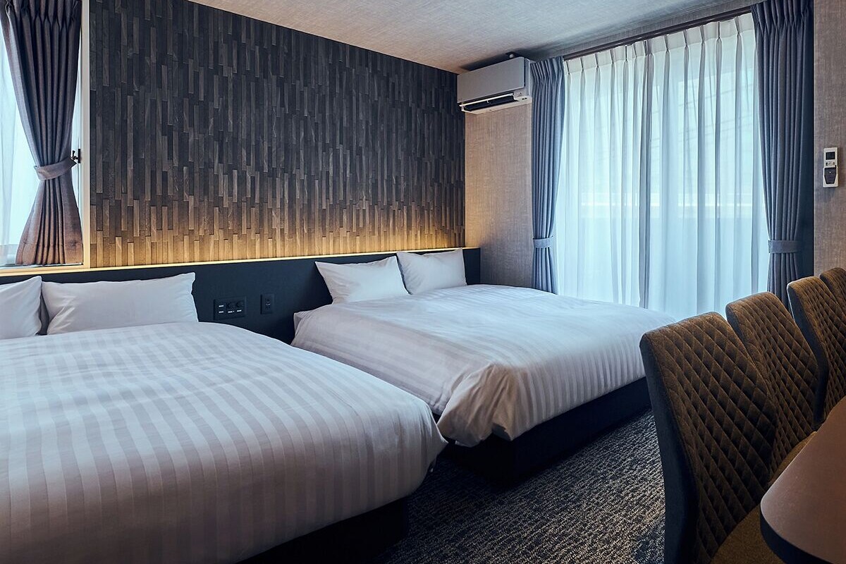Habitación de hotel con dos camas, cabecera, mesita de noche, una silla y una ventana con cortinas.