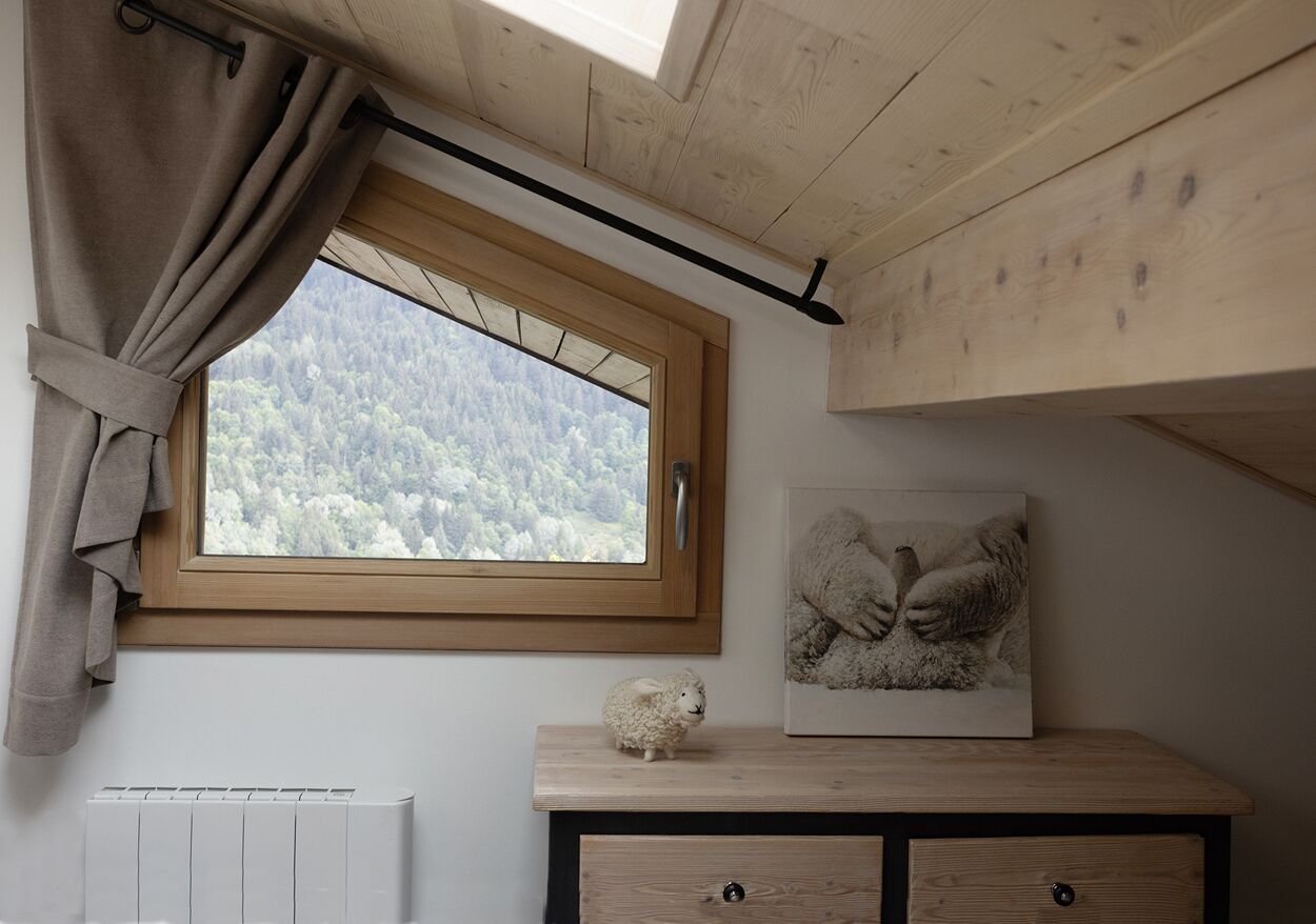 Una habitación con ventana que da a un bosque, un radiador, un armario de madera y un cuadro con ovejas.