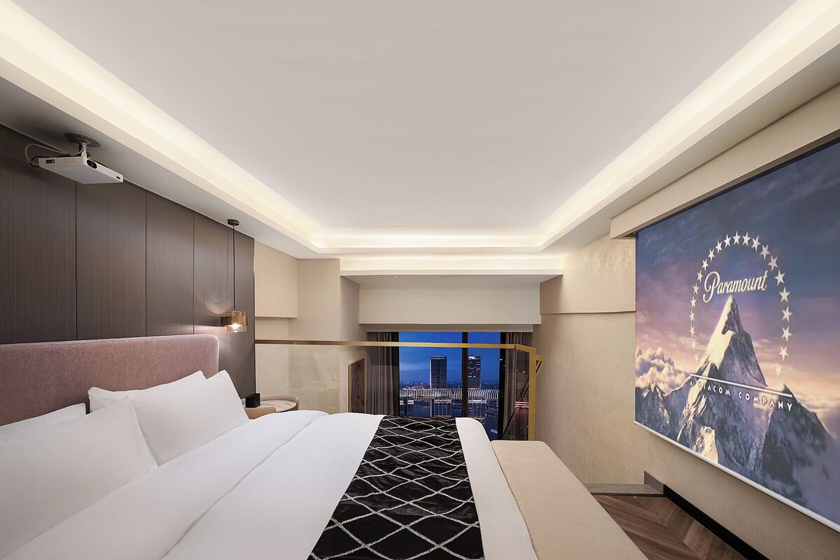 A modern hotel room with a large bed, a city view, and a Paramount movie poster on the wall.