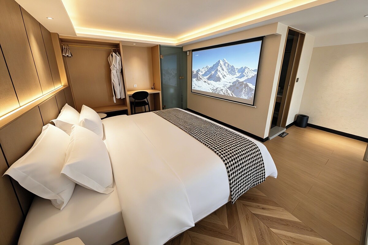 A modern hotel room with a large bed, a desk, a wardrobe, and a large window with a mountain view.