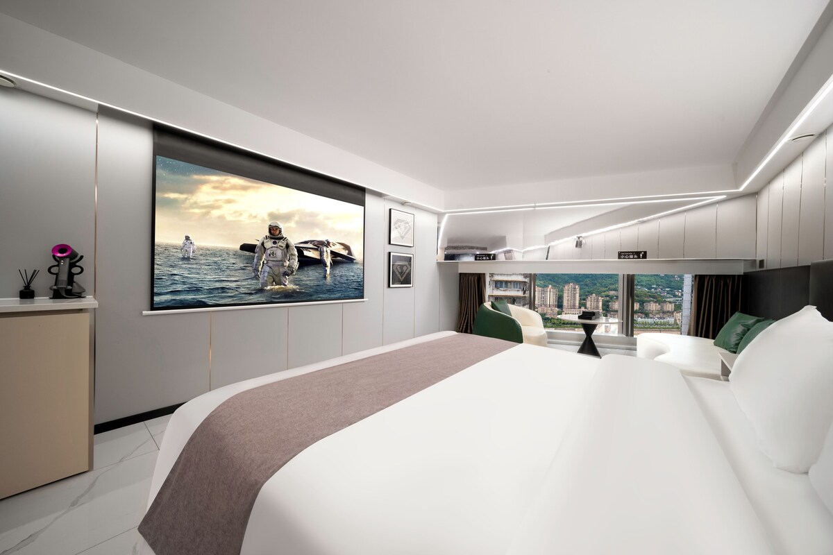 A modern hotel room with a large bed, a desk, a view of the city, and a poster of astronauts on the wall.