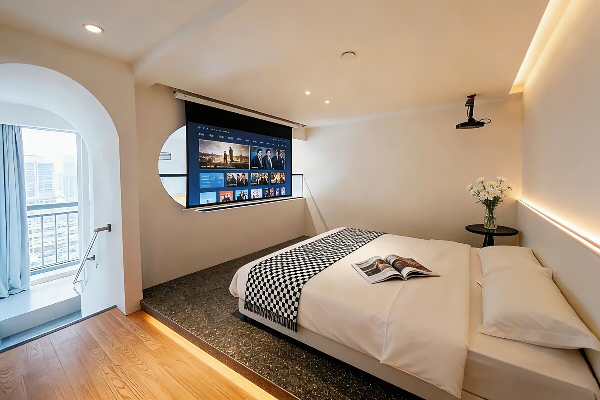 A modern hotel room with a bed, a TV mounted on the wall, a small table with a vase of flowers, and a view of the city through a window.