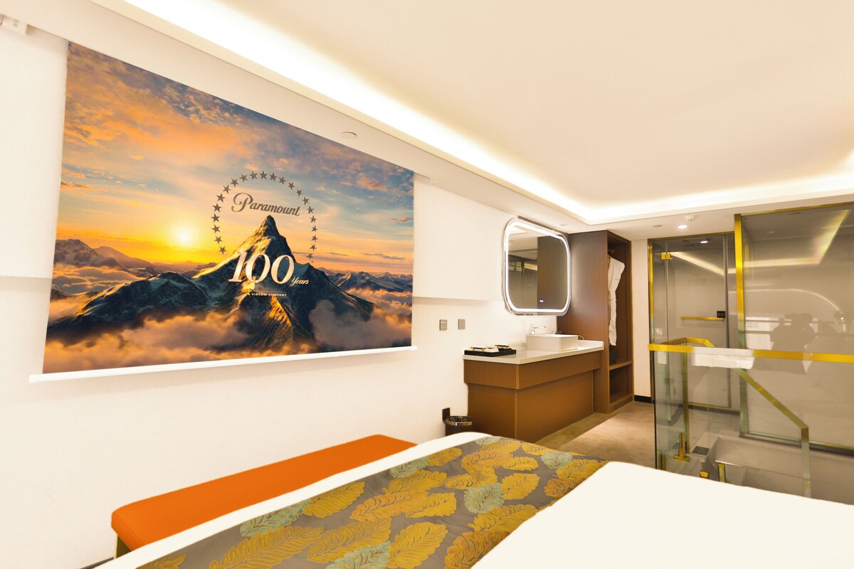 A hotel room with a large wall mural, a modern bathroom, and a bed with a patterned bedspread.